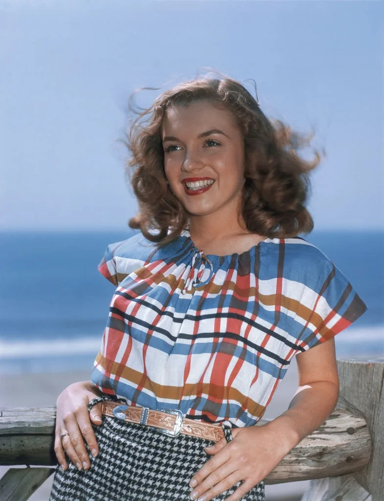 Person smiling near the ocean, wearing a vintage plaid top and gingham skirt, standing at a wooden railing