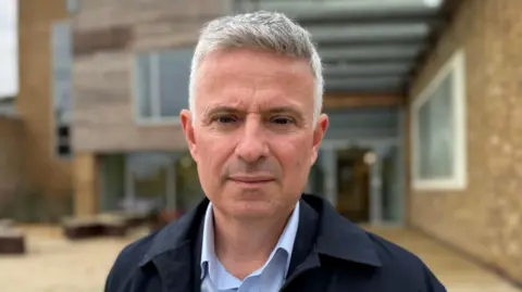 Stuart Hubbard/BBC Paul Scott has short grey hair and is shaven. He wears a light blue shirt and navy blue jacket, and is standing outside a modern building.