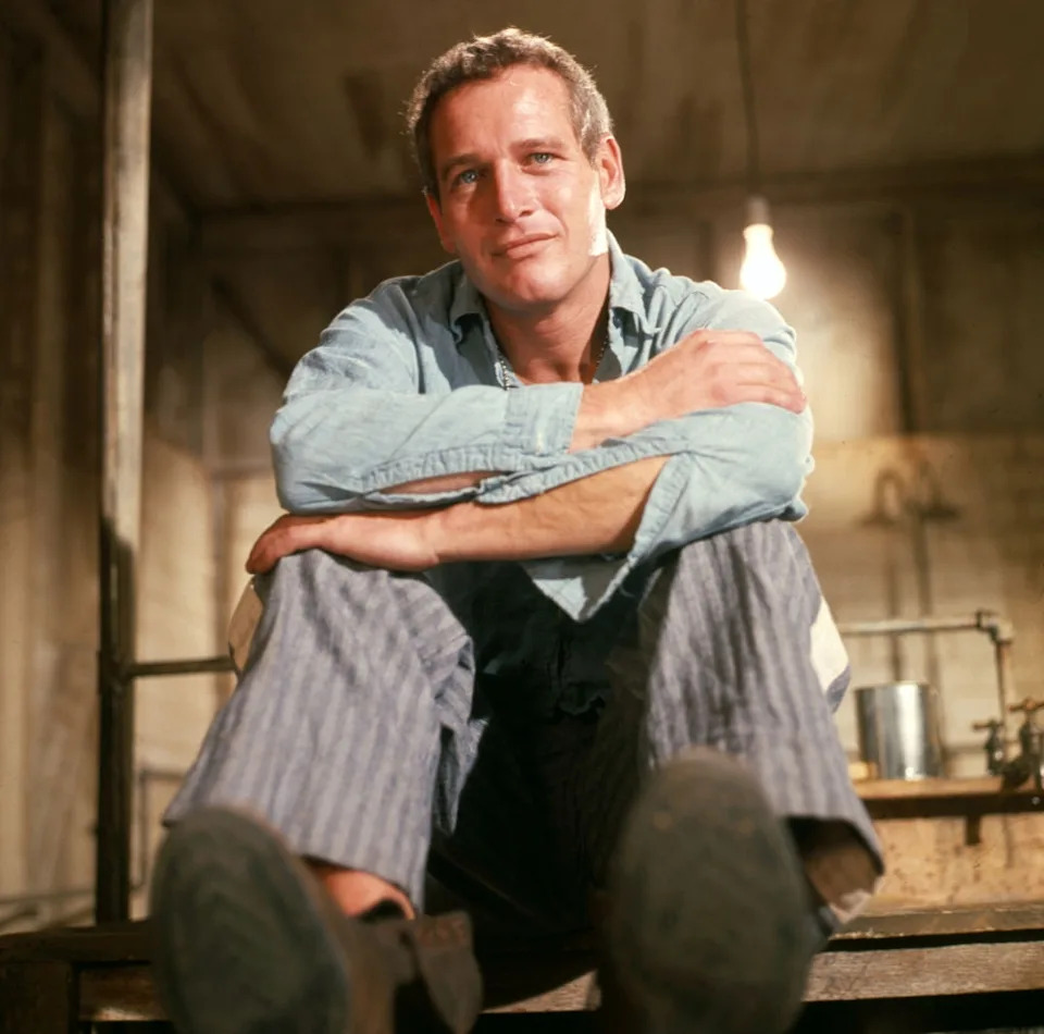 Person sitting casually indoors on a platform, wearing a light shirt and striped pants, gazing directly at the camera with a thoughtful expression