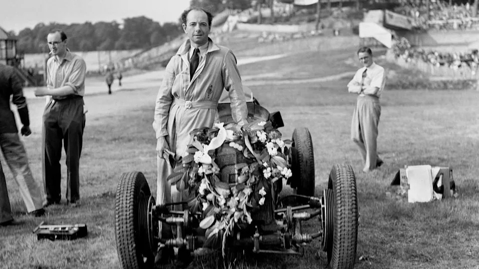 A black and white photo of a man wearing a one-piece racing suit as he leans on a racing car next to him that has a flower wreath on the front of it. A few men can be seen stood around him on a field.