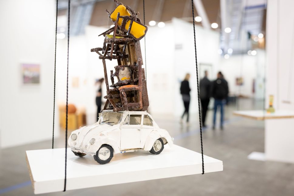 A small white model car with a precarious stack of miniature furniture and yellow cushions on its roof is displayed on a suspended platform at Artissima, with fair visitors blurred in the background.