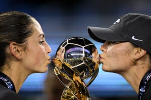 Two women kissing a shiny soccer trophy, celebrating victory, with reflections of a stadium in the background.