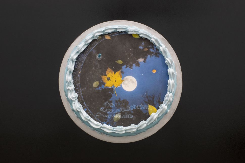 A circular sculptural work resembling a decorated cake shows a printed image of a moonlit sky with a yellow leaf and text in Italian, referencing celestial and earthly cycles.