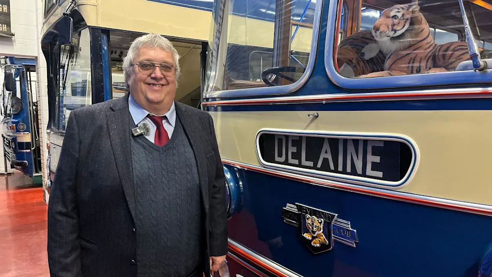 A man with short grey hair and glasses wearing a navy blazer, knitted sweater, red tie and blue shirt is stood next to a vintage yellow and blue bus which says 'Delaine' on it and has a toy tiger in the windscreen.