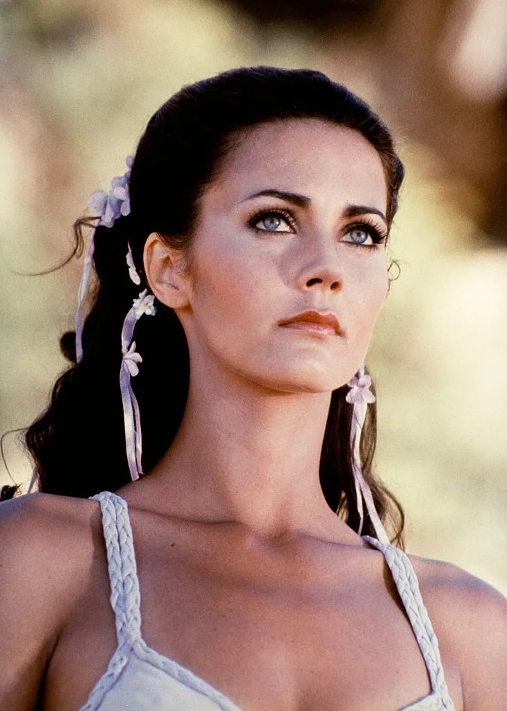 Person with long dark hair adorned with flowers gazes upwards. They wear a braided strap garment, exuding an ethereal, classic style