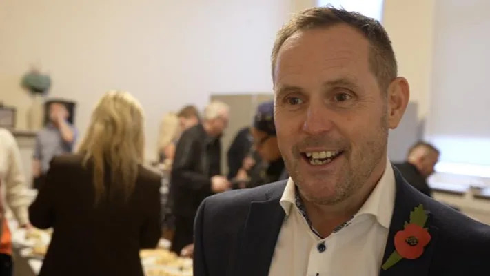 Chris Louis smiles widely at the camera inside a large room where people mingle behind him. He wears a navy suit with a white shirt underneath. He also wears a red poppy in his chest. He has short brown hair and some stubble. 
