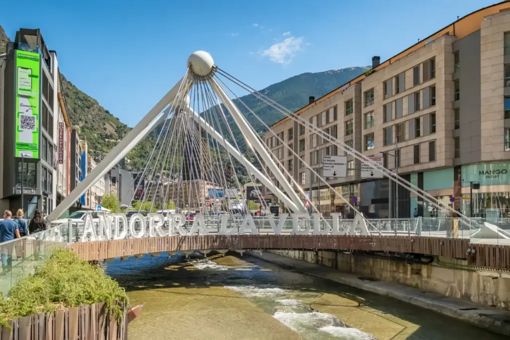 Pedestrian bridge with “Andorra la Vella” sign spanning a river in the city center, surrounded by modern buildings and mountain scenery.
