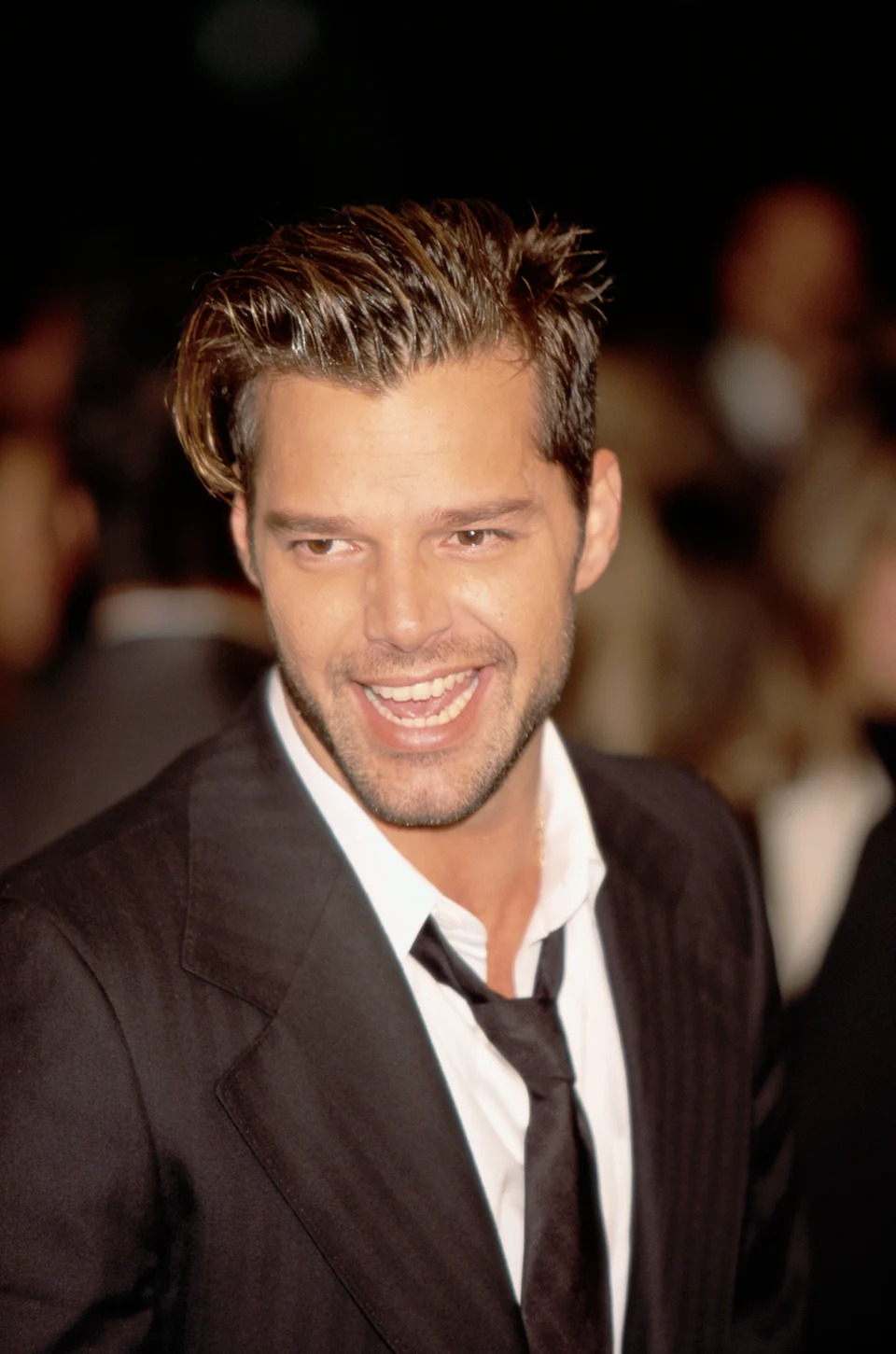A smiling man in a suit and tie on a red carpet event. His hair is styled neatly back, and he exudes a confident and charismatic presence