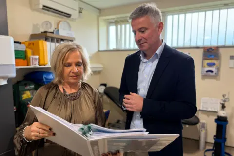 Stuart Hubbard/BBC Paul Scott standing beside Martine Munby at Brockfield House. He is standing in the office with a medical bed in the background. Ms Munby is holding a white folder in front of her and is looking at it. Mr Scott is looking over Martine's shoulder a the contents of the folder. There is a high window and you can see an air conditioning unit, a clock and shelves in the background