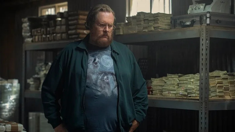Steve Agee as John Economos looks serious in front of a lot of money, wearing a glorious three raccoon moon shirt