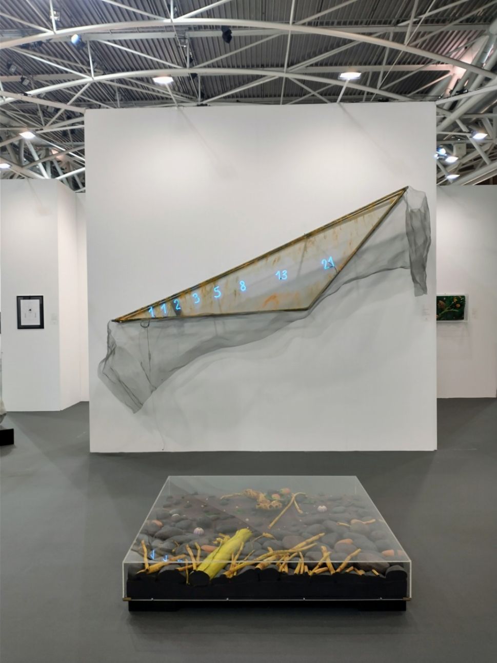 An installation view at Artissima shows a mixed-media artwork with a translucent net-like fabric stretched across a wall displaying glowing numbers, and a glass-covered box on the floor containing stones and yellow plant-like objects.