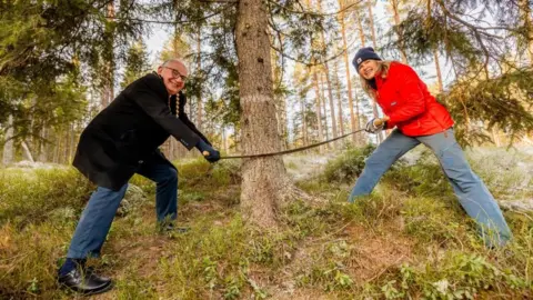 Sturlason/Oslo kommune The two mayors stand on either side of a tree trunk with a two-man saw. They look at the camera and smile with their legs braced. Mayor Anne Lindboe wears a hat and orange coat and Mayor Paul Dimoldenberg wears a black coat with blue jeans.