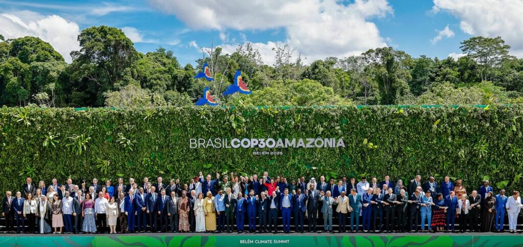 Hundreds of people lined up on a sunny day in front of a trimmed flat section of huge, tall bushes with green trees visible in the background. A logo on the bushes says: