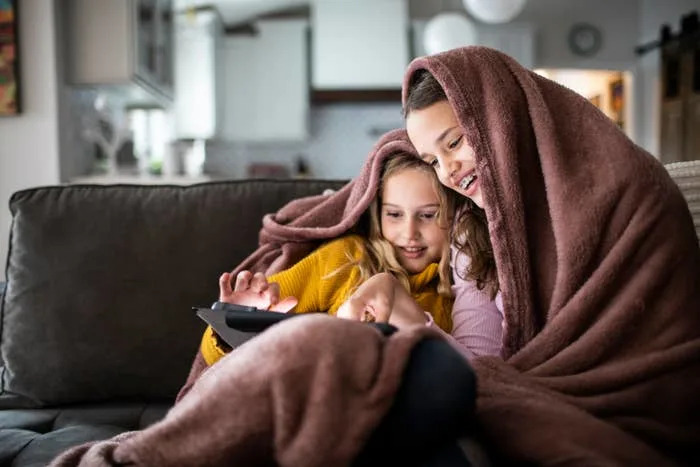 Two children are sitting on a couch, cozy under a blanket, intently watching a tablet together