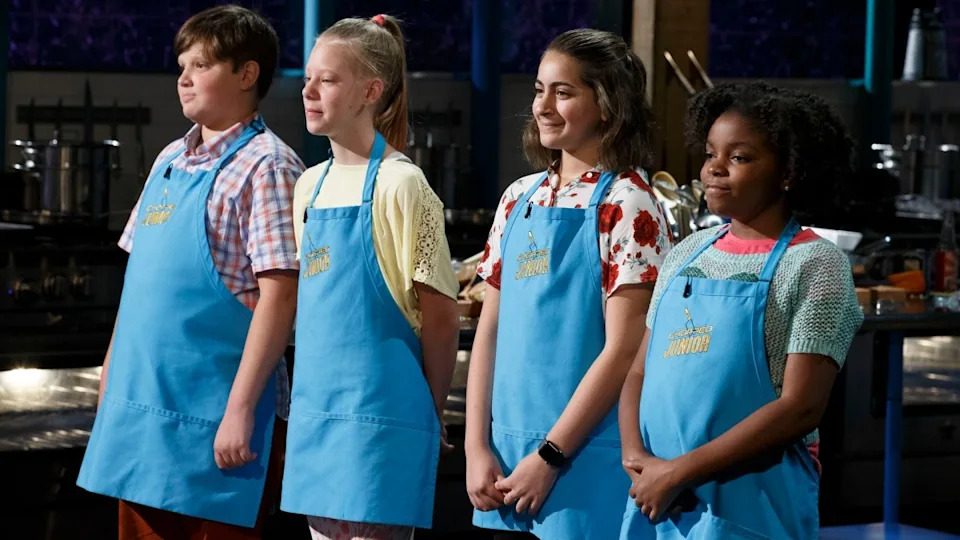Four children stand in a kitchen, wearing blue aprons over casual clothing, participating in a cooking competition