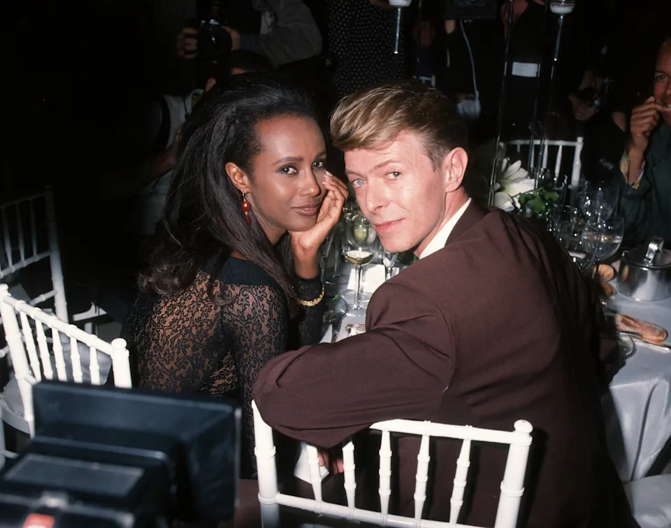Two people at an elegant event, seated at a table. One wears a lace outfit, the other a suit. They glance back at the camera with a slight smile