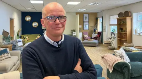 BBC Simon Robinson stands in front of his sofa showroom. He has thick-rimmed black glasses, and wears a blue striped shirt and a blue jumper. In the background, a variety of sofas and staged living room set-ups can be seen.