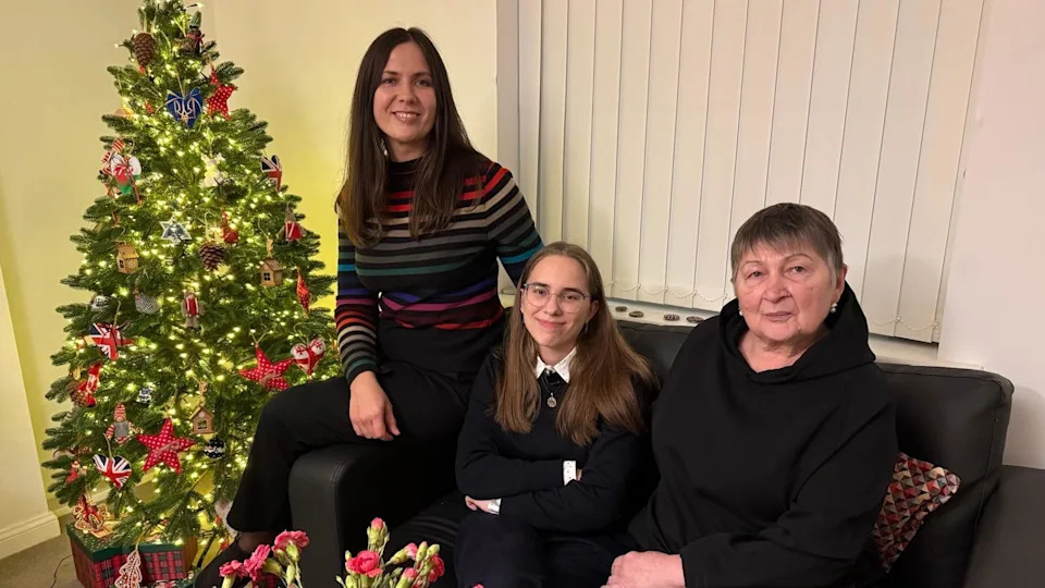 Olena, Kateryna and the 16 year old's grandmother - also called Kateryna all sit on a sofa next to a Christmas tree. The mother wears a stripy jumper, with Kateryna in school uniform and her grandmother dressed in black