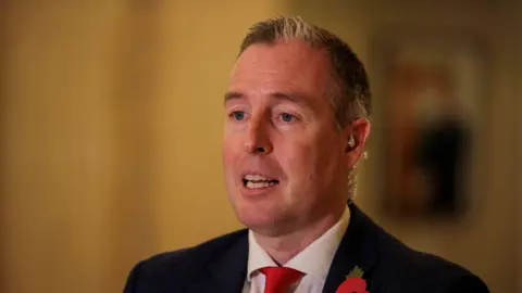 PA Media Paul Givan speaking. He is wearing a black suit jacket, white shirt and red tie.