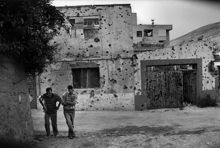 Two people walk down a street in front of a damaged building.