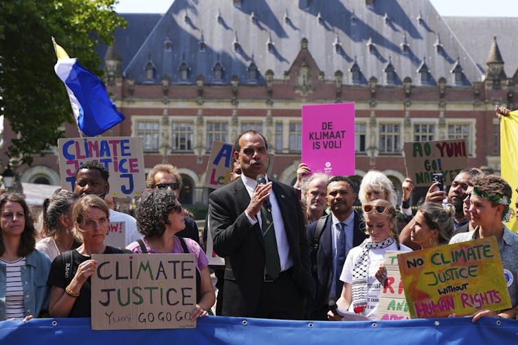 A man in a suit (Vanuatu's minister for climate change Ralph Regenvanu) speaks into a microphone with demonstrators behind.
