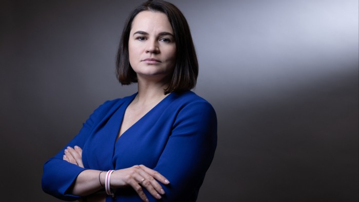 Sviatlana Tsikhanouskaya poses with arms crossed, wearing a blue top, against a neutral background.