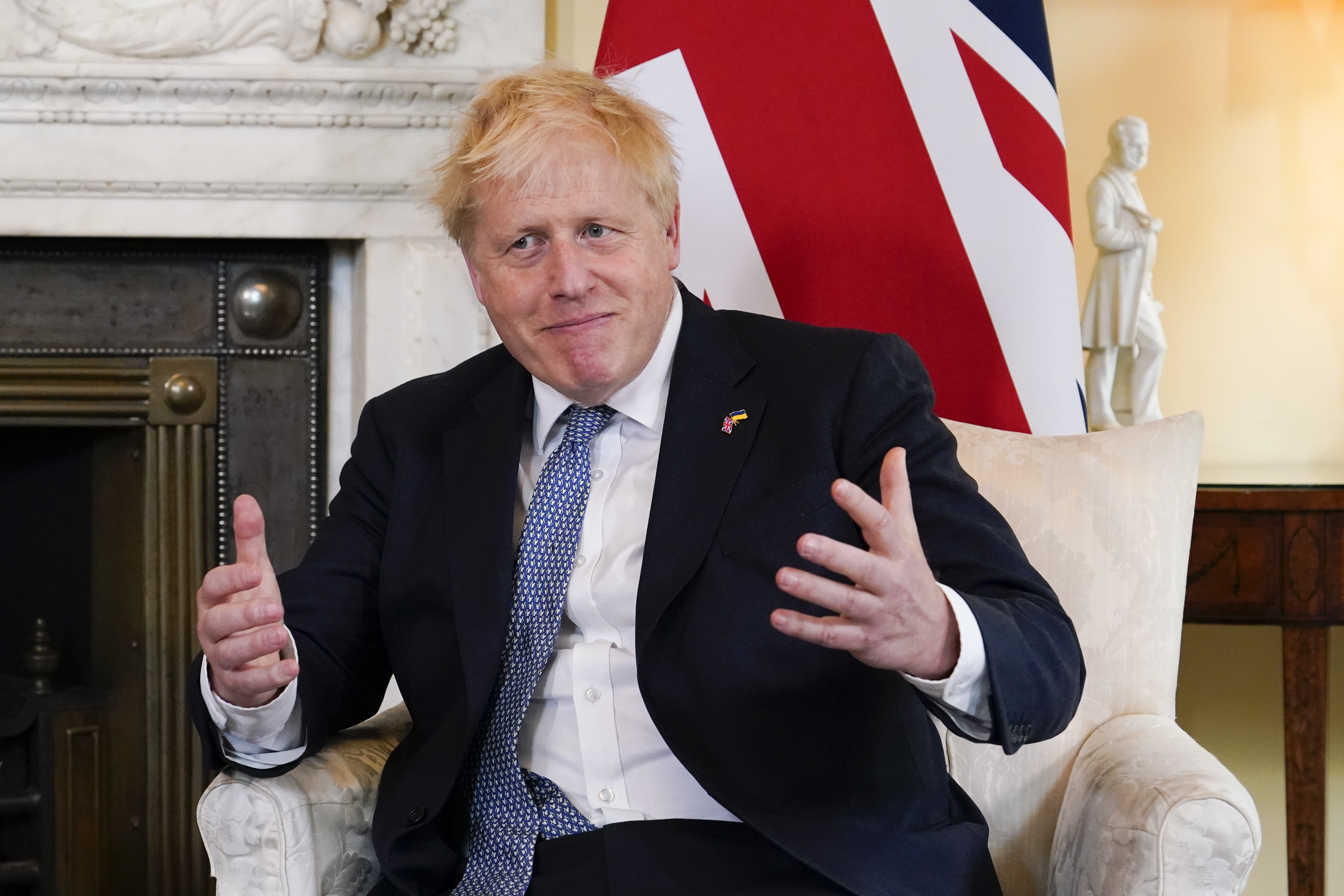 Britain's Prime Minister Boris Johnson gestures while sitting in a white chair, with a Union Jack flag behind him.