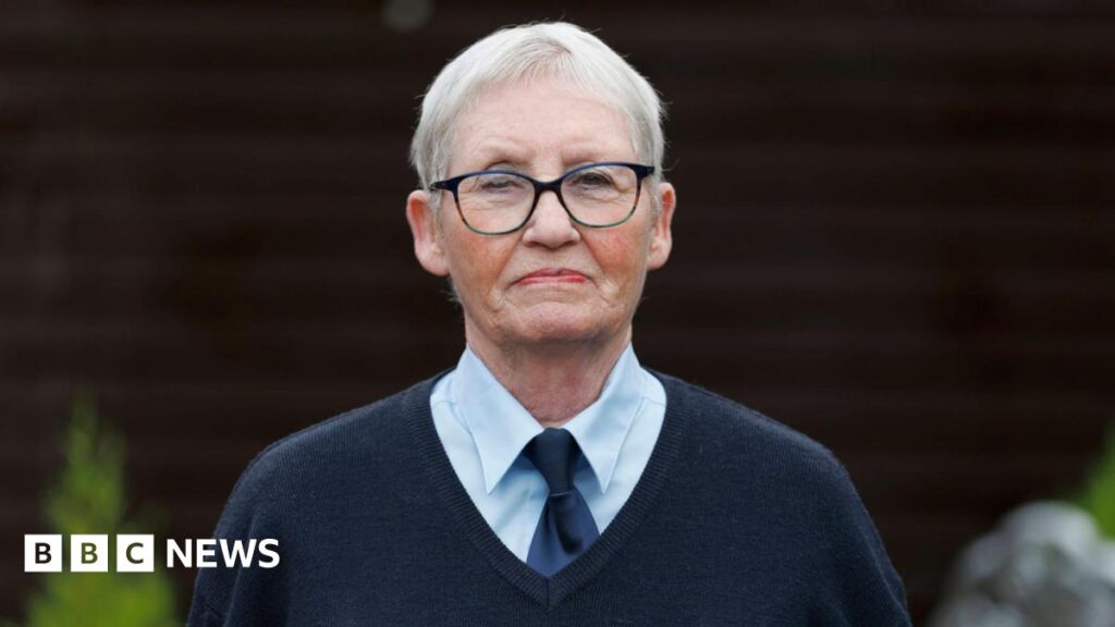 Elizabeth Hamilton has short grey hair and black-framed glasses and is wearing a navy v-neck jumper and tie