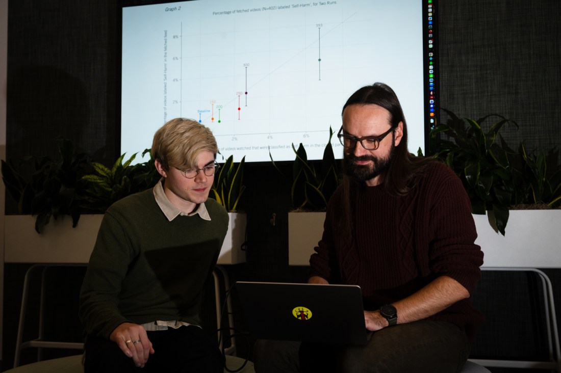 Levi Kaplan (left) and Piotr Sapiezynski (right) looking at a laptop together. In the background, a large screen is displaying a graph titled 'Percentage of fetched videos (N=402) labeled 'Self-Harm,' for Two Runs'. 