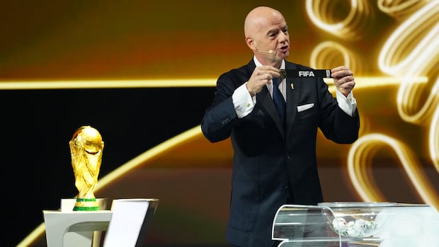 A man in a black suit holds up a black piece of paper that reads 'FIFA' while on stage next to a trophy.