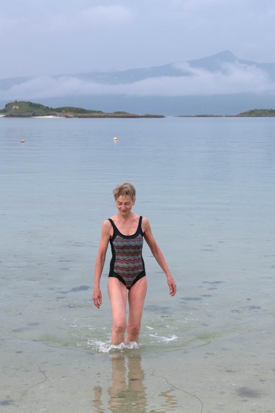 A color photograph of a woman walking through shallow water in a one piece bathing suit