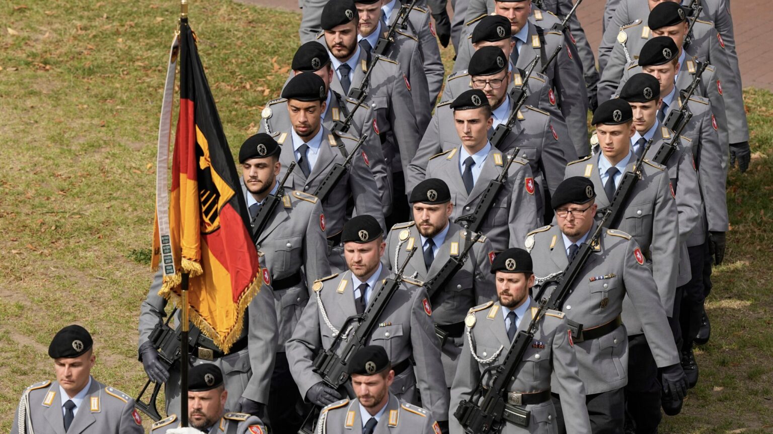 A group of uniformed soldiers marching in formation, wearing gray uniforms and black berets, and carrying rifles. One soldier holds a black, red, and gold flag with an emblem. The soldiers are walking on a grassy area, with fallen leaves visible.