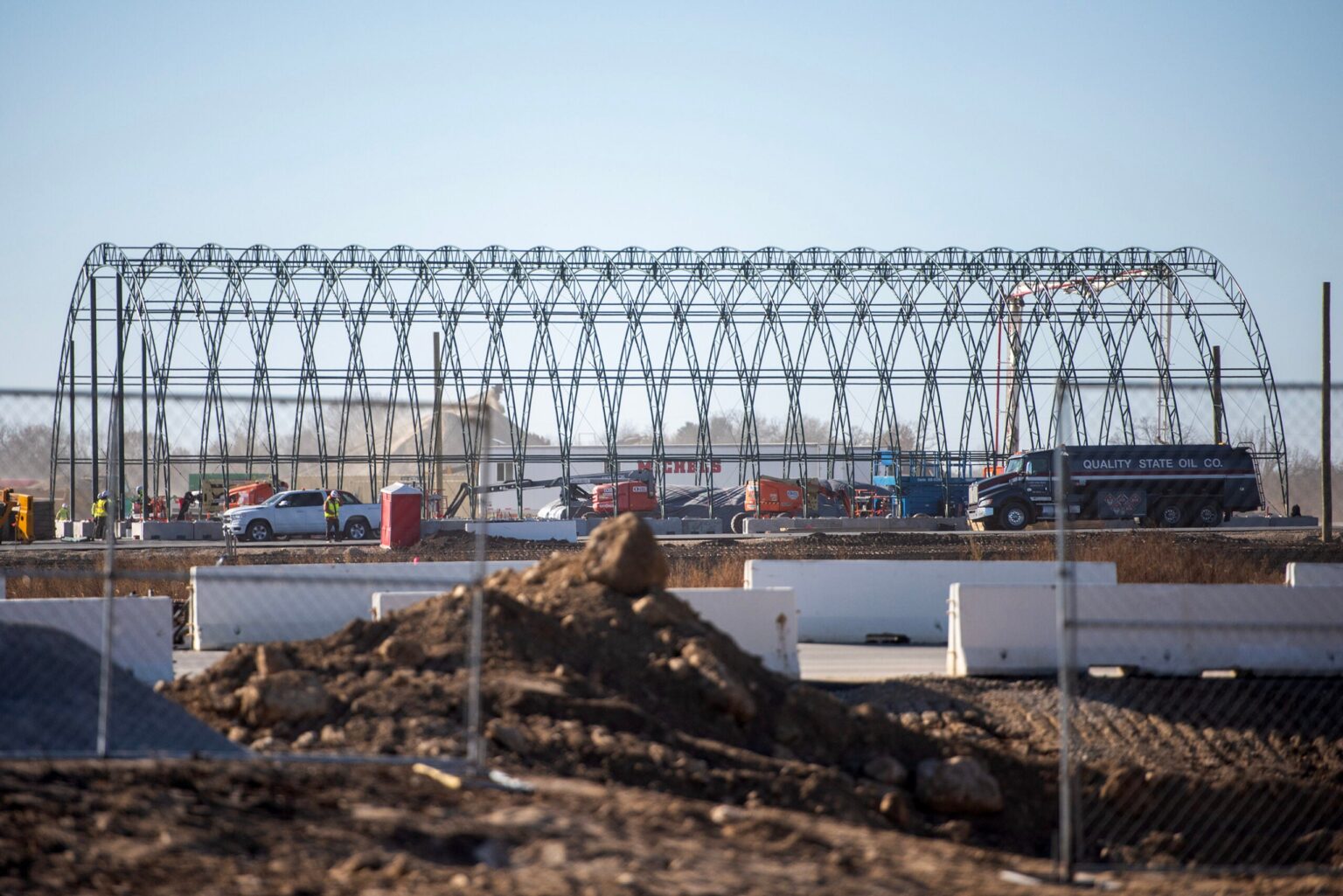 A construction site with a large metal frame structure, vehicles, and equipment is visible behind a chain-link fence on a clear day.
