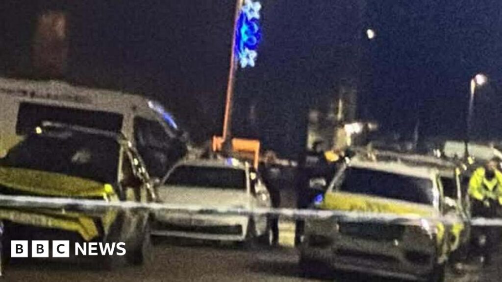 Several police cars and officers on a street in the dark, behind police tape.