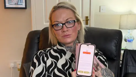 BBC/JIM SCOTT Samantha Fisher wearing black-rimmed glasses and a black and white patterned shirt, is sitting on a black recliner sofa and is holding up her pink and black phone. On the screen is an email from Royal Mail, showing the logo at the top, followed by text which apologises for the ongoing delays she is facing. In the background is a white door, a lamp and a double plug socket.
