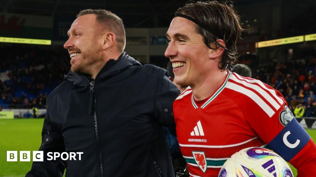 Craig Bellamy and Harry Wilson holding a ball both smile on the field after winning 7-1 against North Macedonia