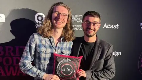 Max Blair Two men stood, smiling at the camera. The man on the left has long brown hair and glasses. He is wearing a chequered shirt. The man on the right has short dark hair and glasses. He is wearing a dark top and a grey blazer jacket. They are holding a square cushion with 'Last Dance' written on it. The cushion is black and red with white writing. 
