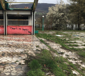 The picture shows a garden. On the right is a grassy area with scattered flat stones. On the left is a stone terrace adjacent to part of an abandoned house. Written in black at the bottom of the building is: 'There's no happy ending.' The wall is painted pink.