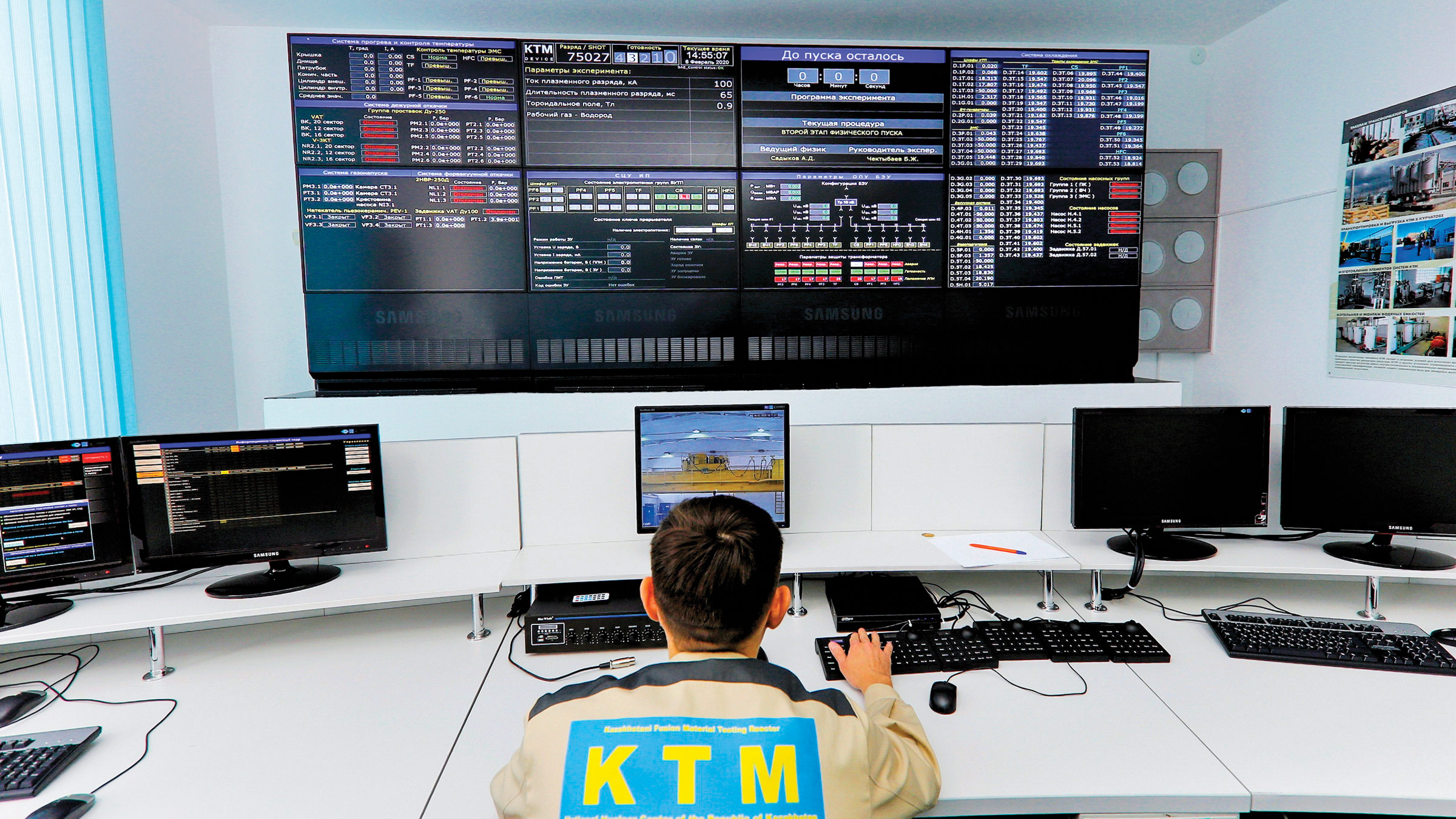 A man wearing a jacket emblazoned with “KTM” on the back monitors a large screen displaying numbers and images.