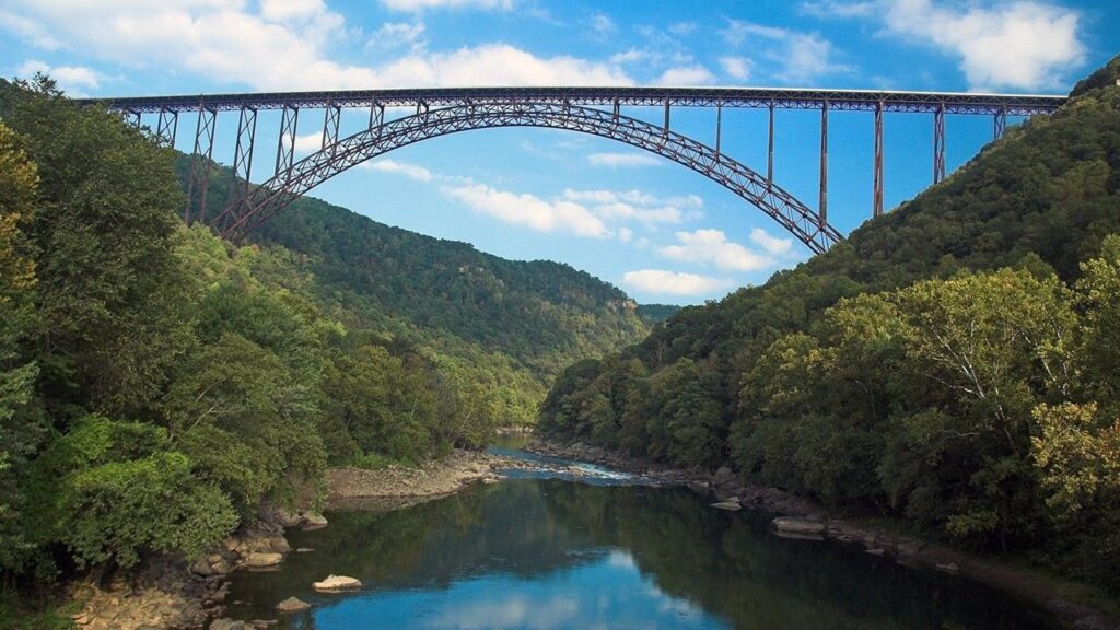 A long bridge supported by metal arch spans a river valley luch with trees in full summer foliage.
