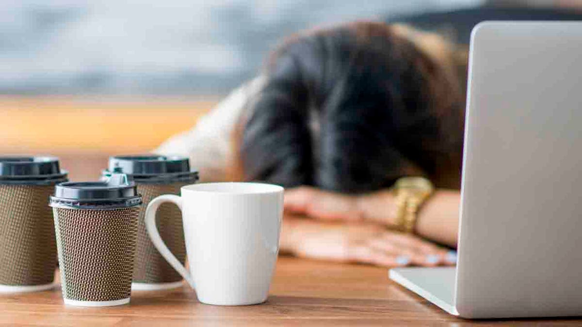 Woman with head down on desk in front of laptop and coffee cups in front of her.
