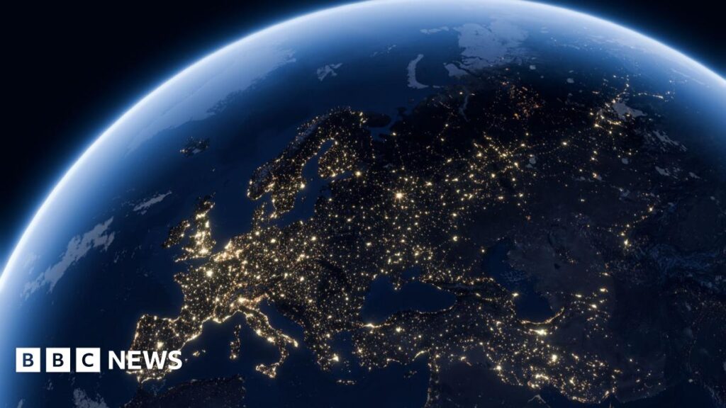 A life-size mock up of the space factory satellite sitting on a workbench inside Space Forge's Cardiff headquarters. It is square-shaped, about the size of a microwave, and is covered in reflective orange foil with various smaller components attached. The workshop in the background has high ceilings and has shelves filled with equipment and storage boxes. A large Union Jack flag hangs on the wall.