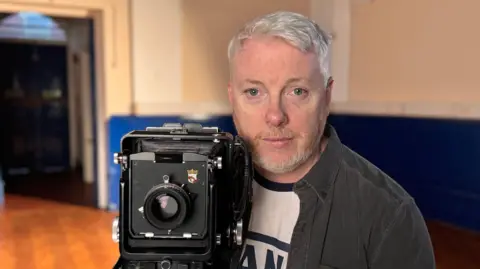 Photographer Stu McKenzie poses with a camera in a sports hall.
