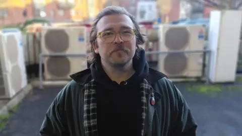 JR McCormick A man with brown hair looks at the camera. He is wearing a jacket and a hoodie, as well as glasses. He is standing outside. There are red-brick buildings in the background. 