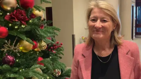 BBC A picture of a lady wearing a black shirt and a pink jacket. She is sat next to a green christmas tree with decorations on it.
