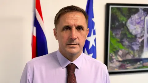 Anes Ceric wears a purple shirt and tie and poses in front of the Union flag and Bosnia-Herzegovina flag.