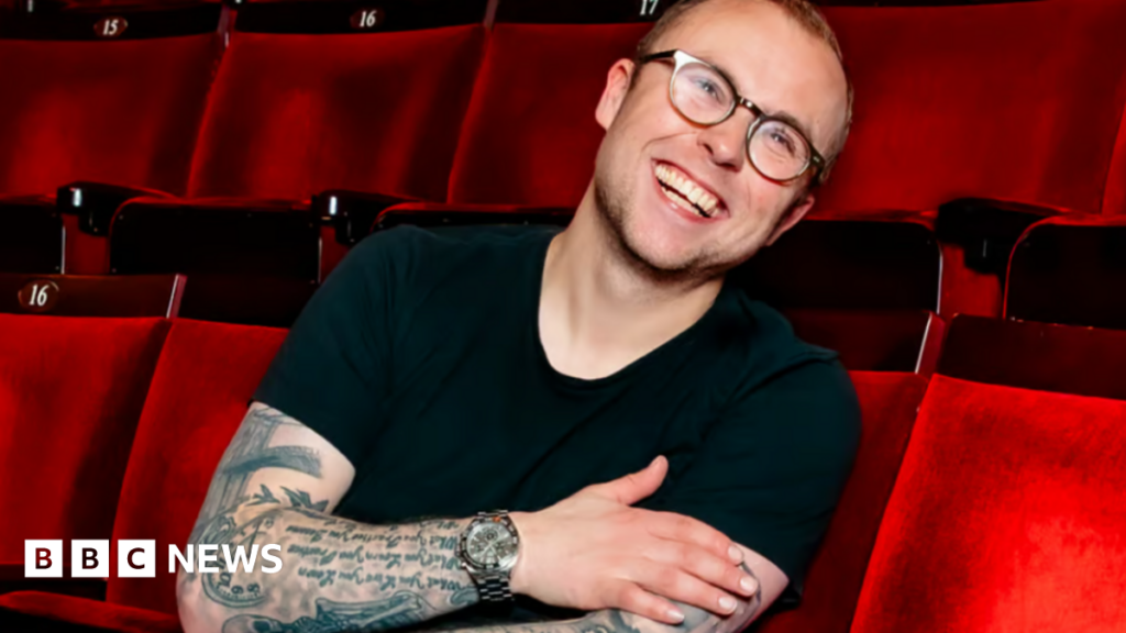 Joe in a black top, wearing glasses, sitting in red theatre/cinema seats