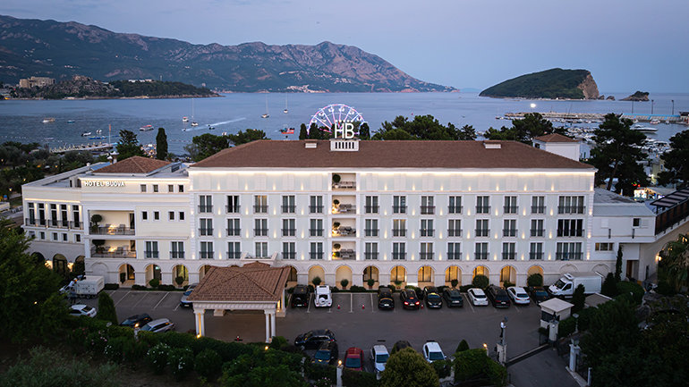 An aerial shot of a large hotel complex situated on a coastline at dusk. The hotel has a multi-story, classic architectural style with many windows and balconies, illuminated by warm lights. In front of the hotel, there's a parking lot with several cars. In the background, a calm sea stretches out, dotted with boats and yachts. Further in the background, there are mountains and an island, all under a soft, twilight sky with hints of purple and blue. A large, illuminated Ferris wheel-like structure with "HB" in the center is visible on top of the hotel.