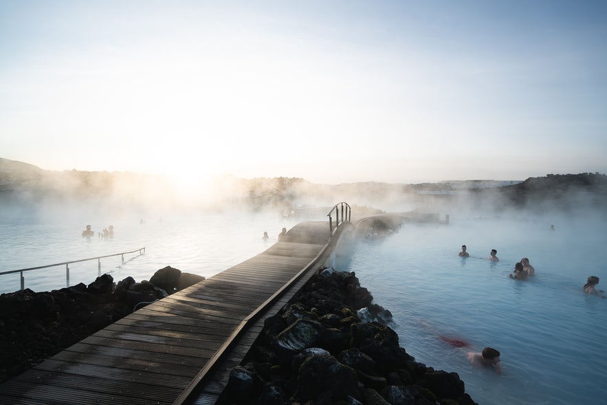 People relaxing in the Blue Lagoon geothermal spa in Iceland, with steaming milky-blue water and wooden bridge at sunrise.