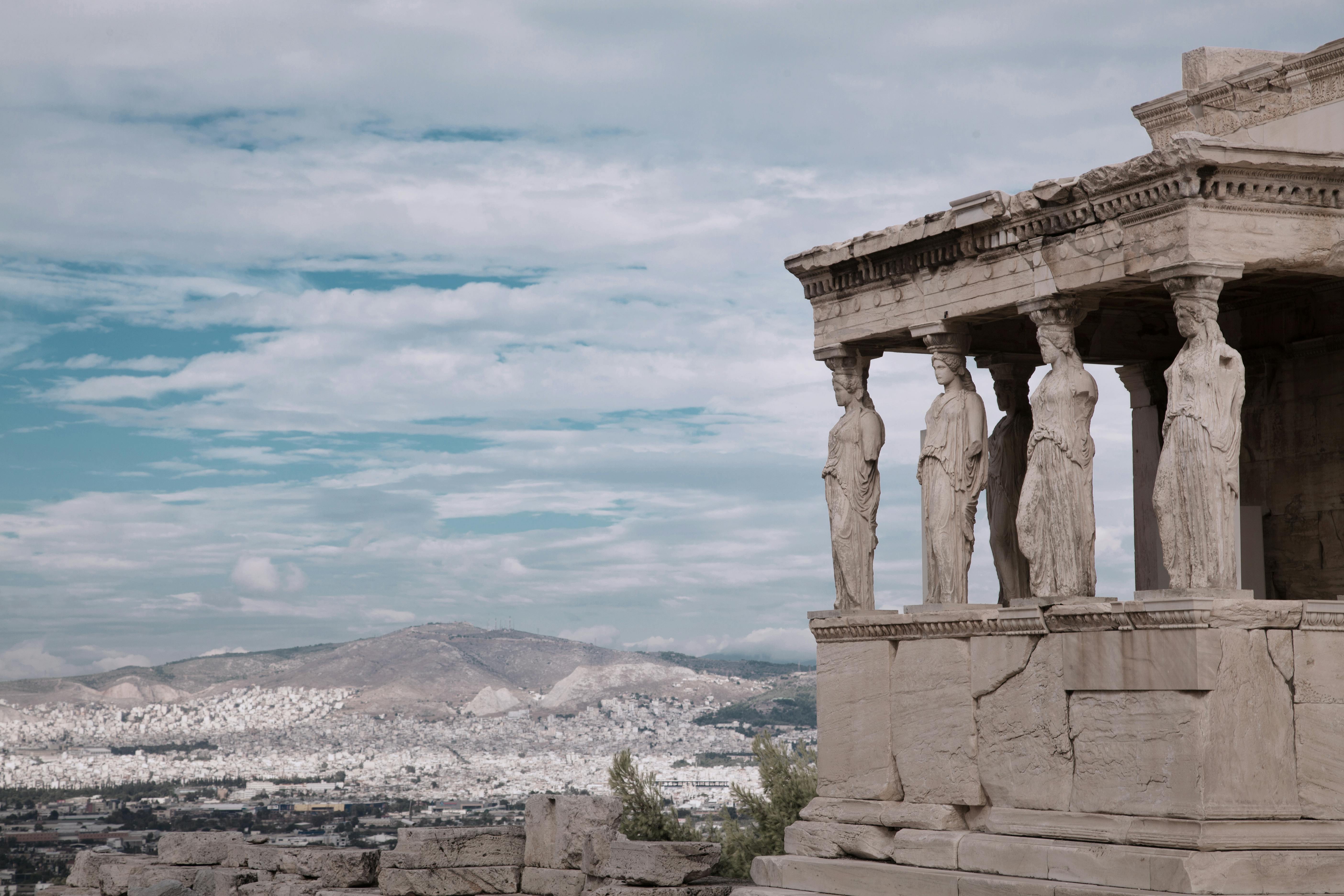 The Erechtheion’s caryatid columns stand on the Acropolis, overlooking Athens and distant mountains under a cloudy sky.